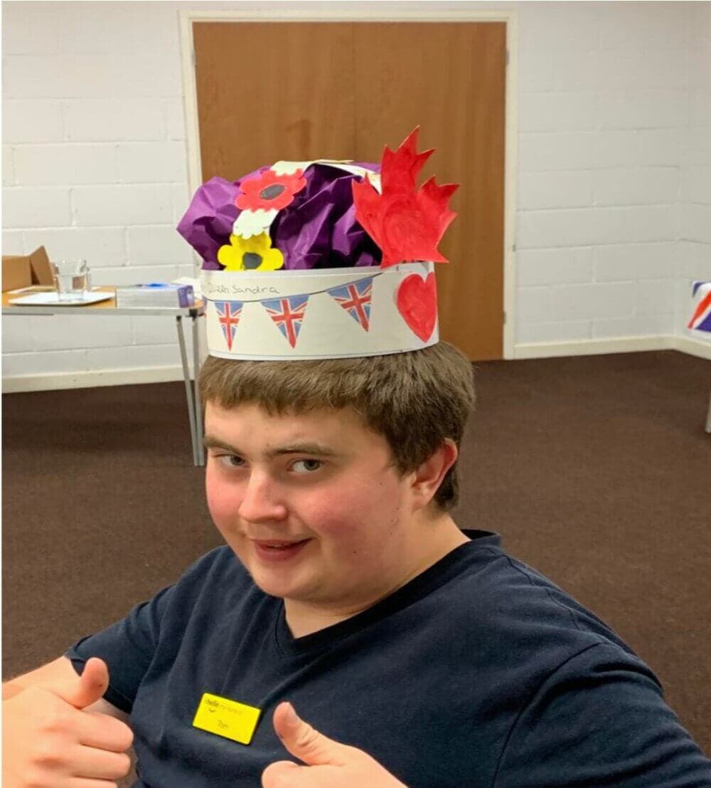Person smiling and giving thumbs up, wearing a handmade crown with British flags, red heart, and colorful paper decorations. - Home Instead