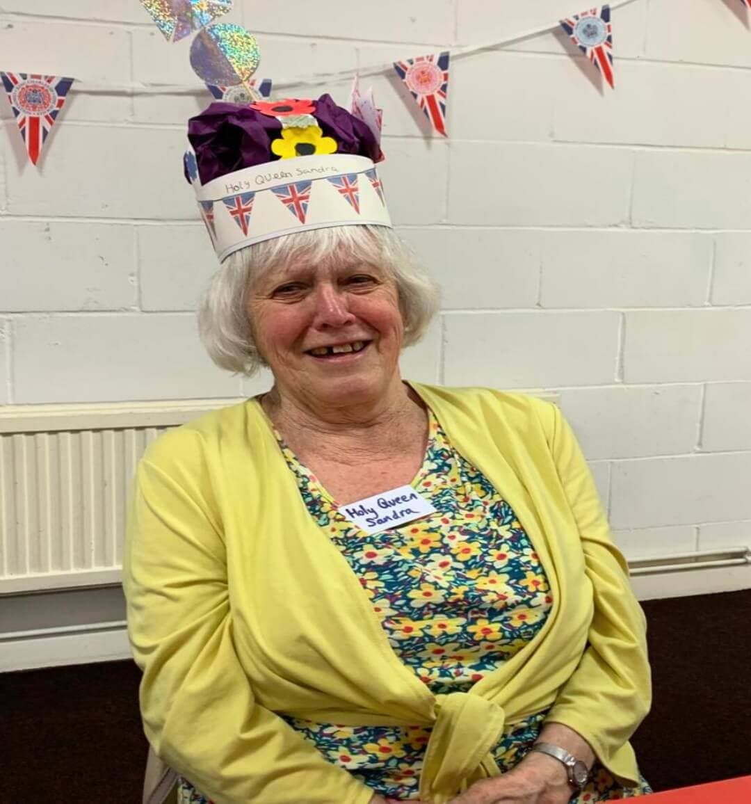 An elderly woman wearing a festive hat and a name tag, sitting in a decorated room with Union Jack bunting. - Home Instead