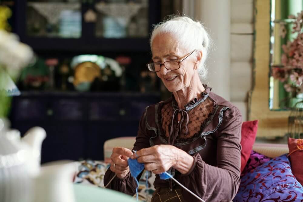 Elderly woman with glasses smiles while knitting a blue piece. She is seated in a cozy, colorful room. - Home Instead