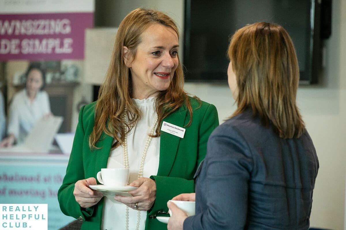 Two women in conversation, each holding a white cup and saucer, during a meeting or networking event. - Home Instead
