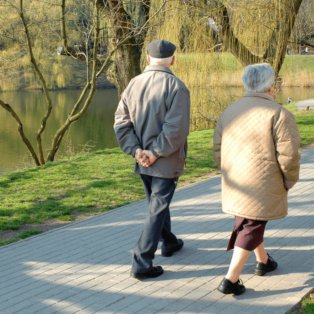 Two elderly people walking side by side on a paved path near a pond, with trees in the background. - Home Instead