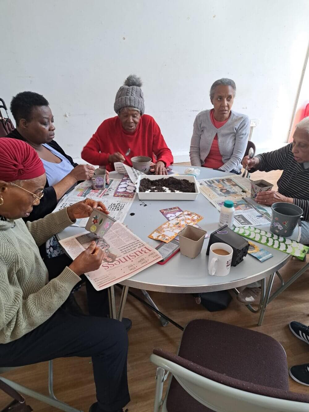 A group of elderly women sits around a table reading and engaging in arts and crafts activities. - Home Instead