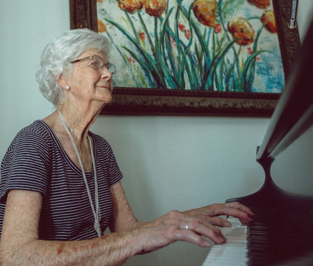 Elderly woman with white hair playing the piano, with a colorful flower painting on the wall behind her. - Home Instead