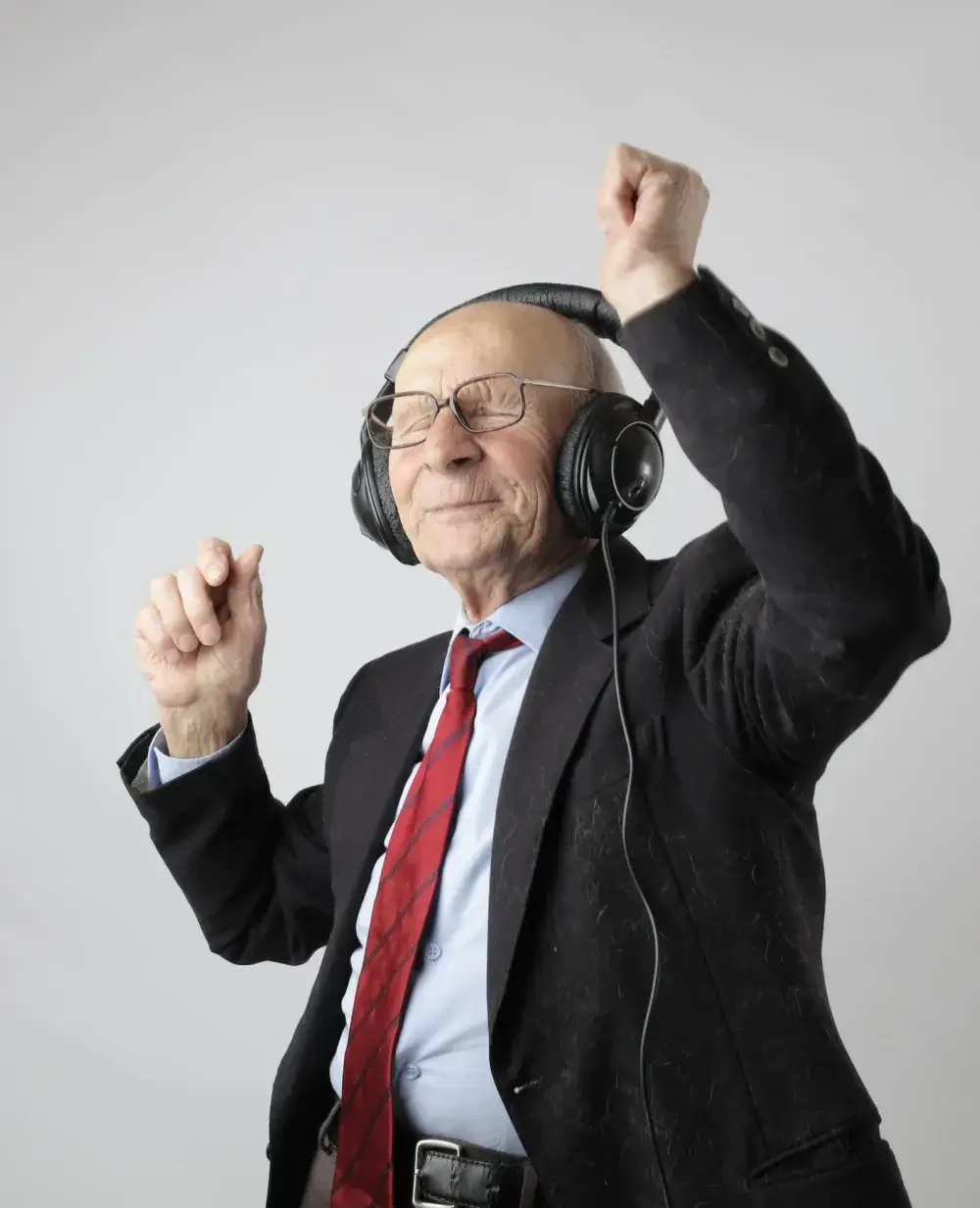 Elderly man in a suit and tie, wearing headphones, dancing with a joyful expression against a plain background. - Home Instead
