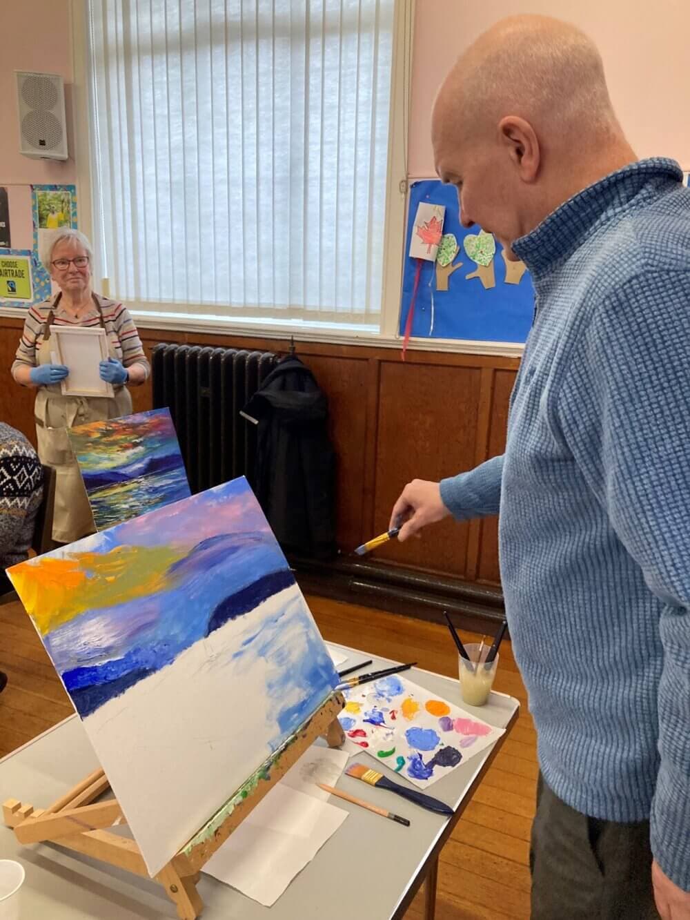 A man paints a colorful landscape on an easel while a woman smiles in the background in a well-lit room. - Home Instead