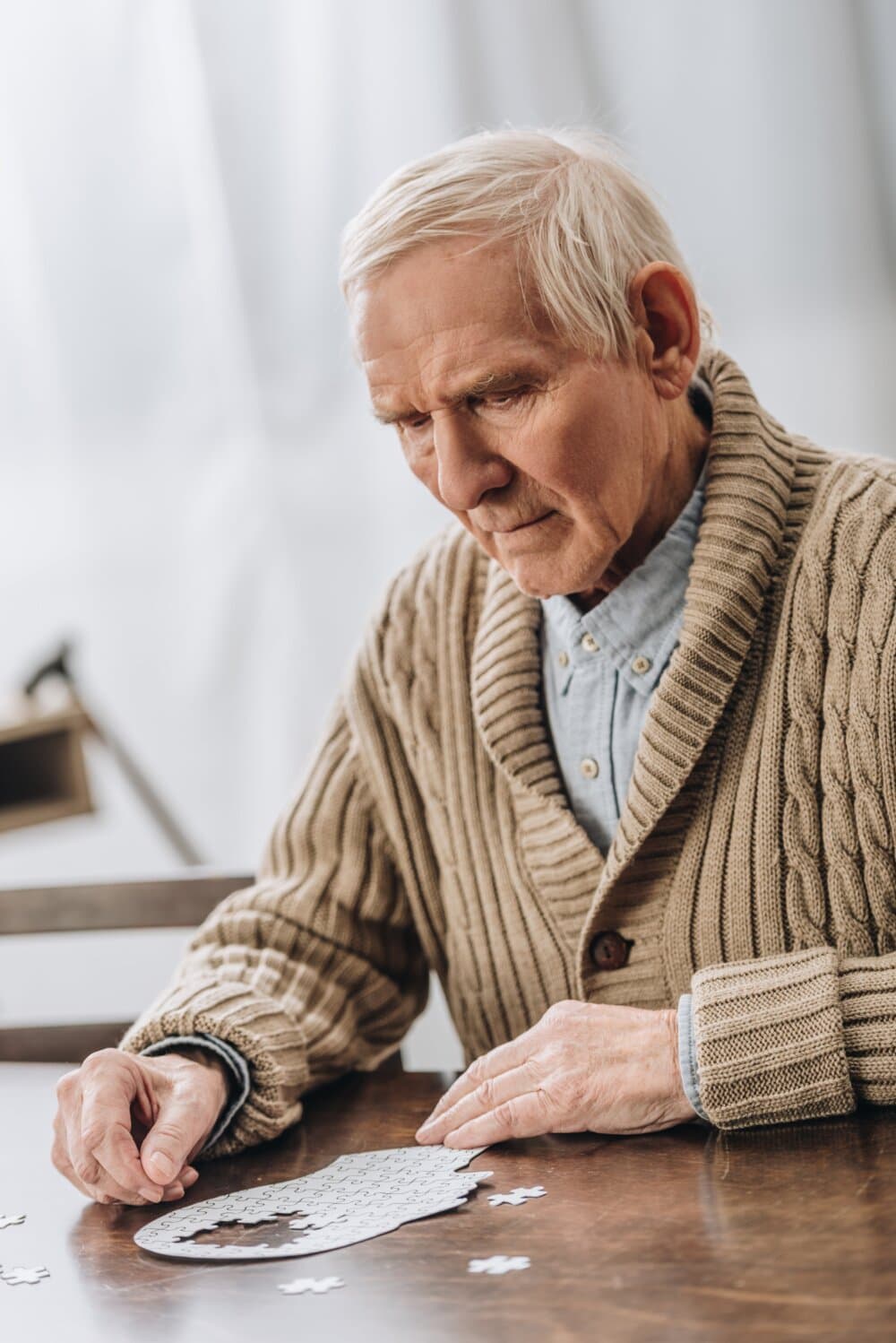 Elderly man in a sweater assembling a jigsaw puzzle at a table, appearing focused and thoughtful. - Home Instead