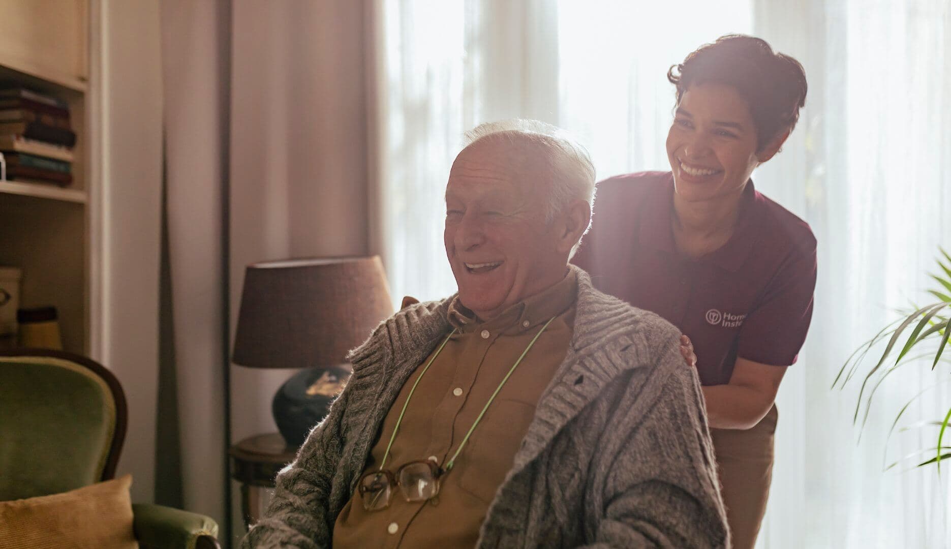 An elderly man smiles while seated indoors, as a caregiver standing beside him smiles warmly. - Home Instead