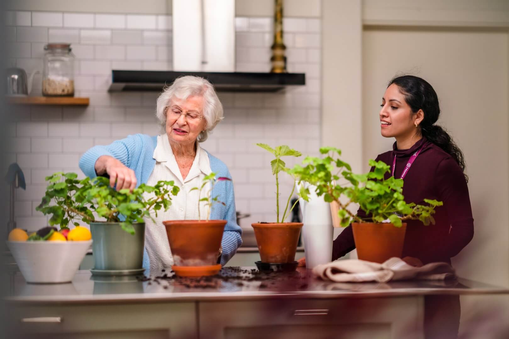 An elderly woman and a young woman tend to potted plants on a kitchen counter with vegetables and containers around. - Home Instead