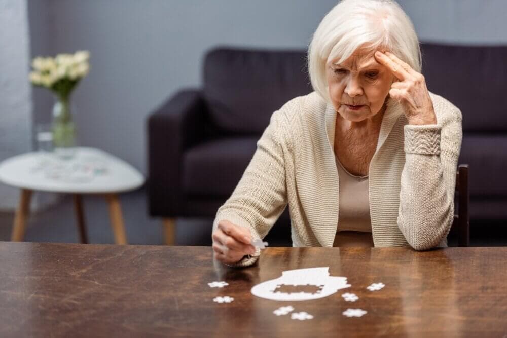An elderly woman concentrating on completing a jigsaw puzzle at a table in a living room. - Home Instead
