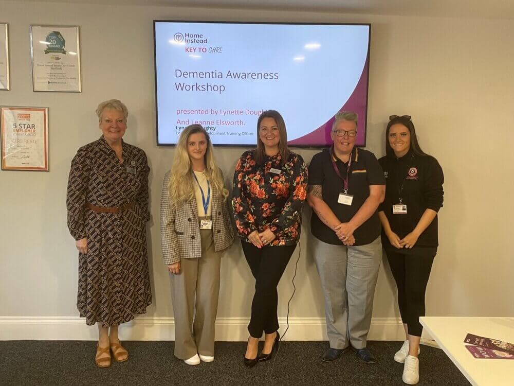 Five people standing in front of a "Dementia Awareness Workshop" presentation screen, smiling at the camera. - Home Instead