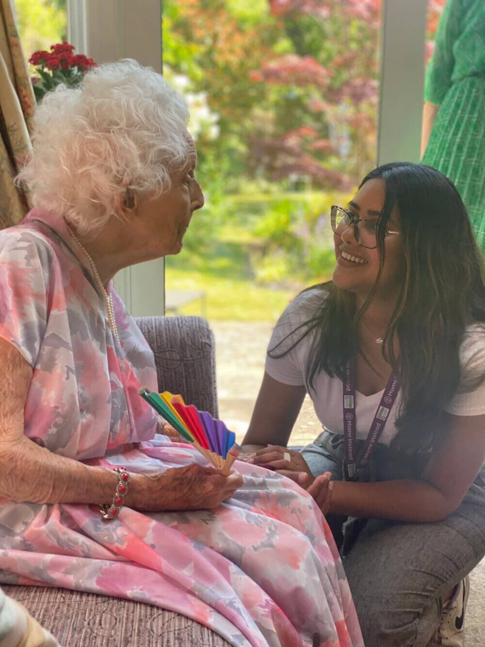 An elderly woman with white hair sits holding colorful papers, chatting with a smiling young woman wearing glasses. - Home Instead
