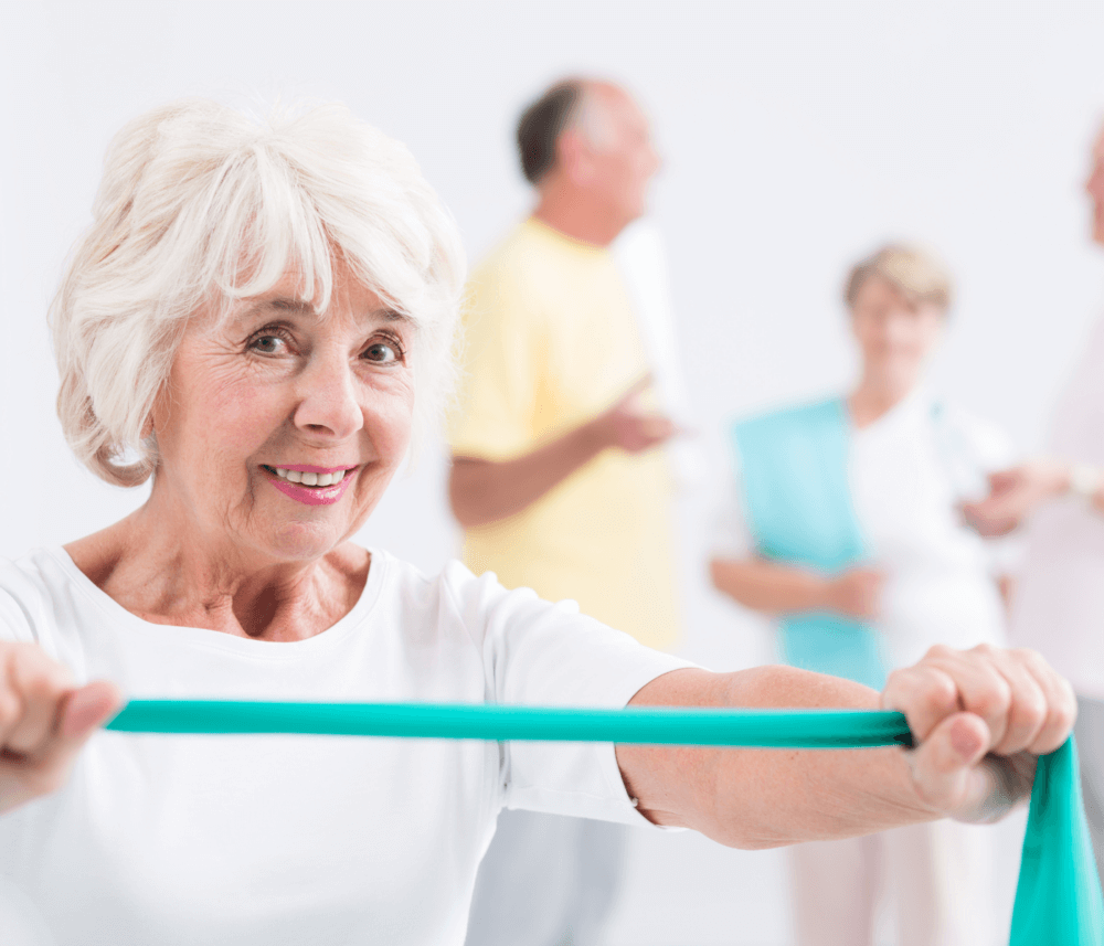 Elderly woman smiling and holding an exercise band, with two men and another woman chatting in the blurred background. - Home Instead