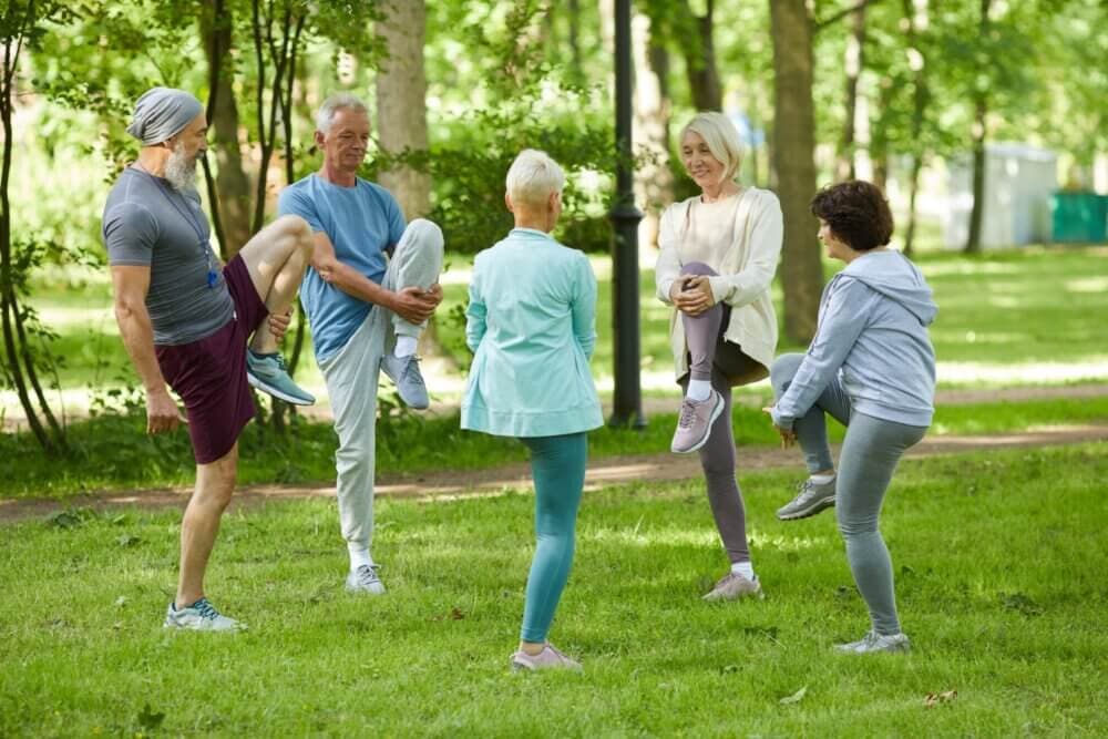 A group of older adults exercises together in a park, lifting their legs and stretching. - Home Instead