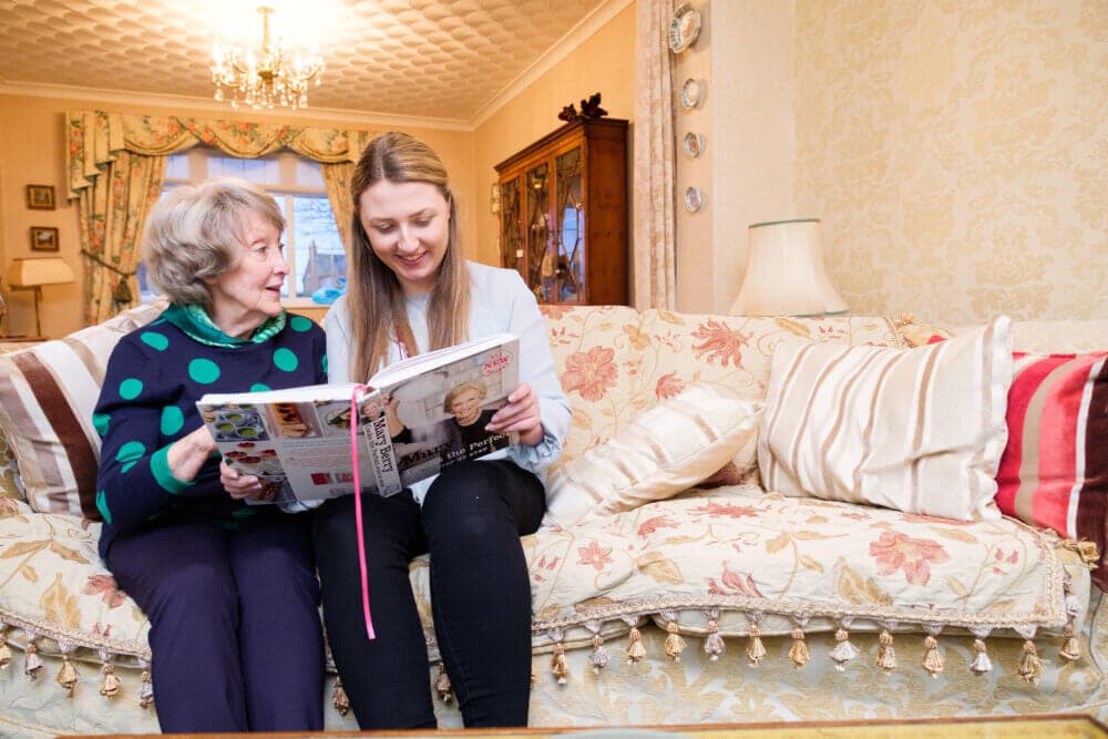 An elderly woman and a young woman share a happy moment on a floral couch while looking at a book together. - Home Instead