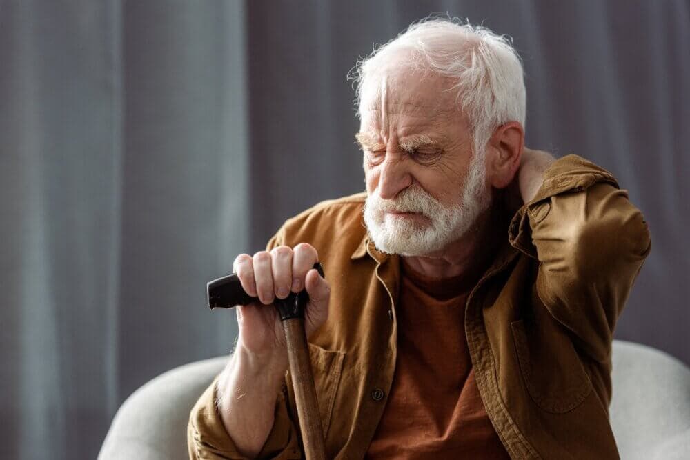 Elderly man with white hair and beard holding a cane, looking pained, and rubbing his neck while sitting. - Home Instead