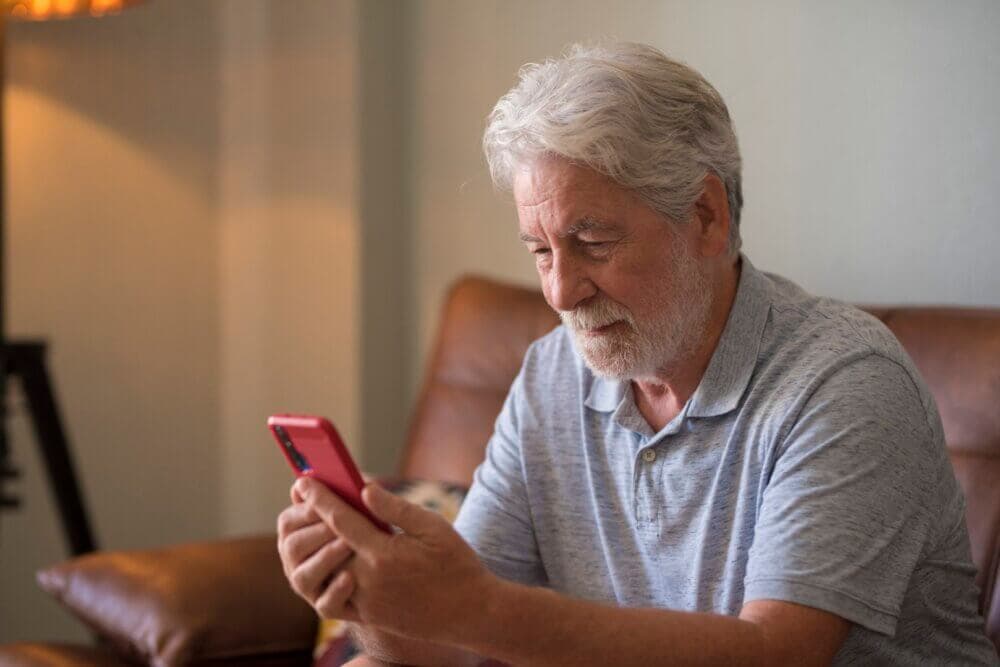 Elderly man with gray hair and beard smiles while looking at his phone, sitting on a brown leather couch. - Home Instead