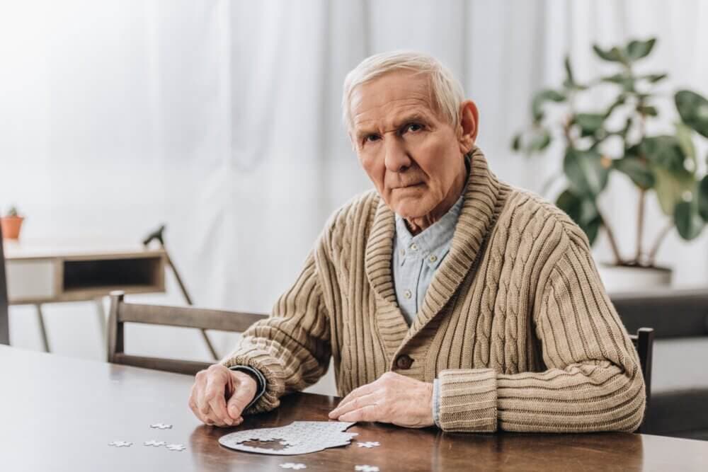 Elderly man in a beige sweater sitting at a table, working on a puzzle, with a pensive expression. - Home Instead