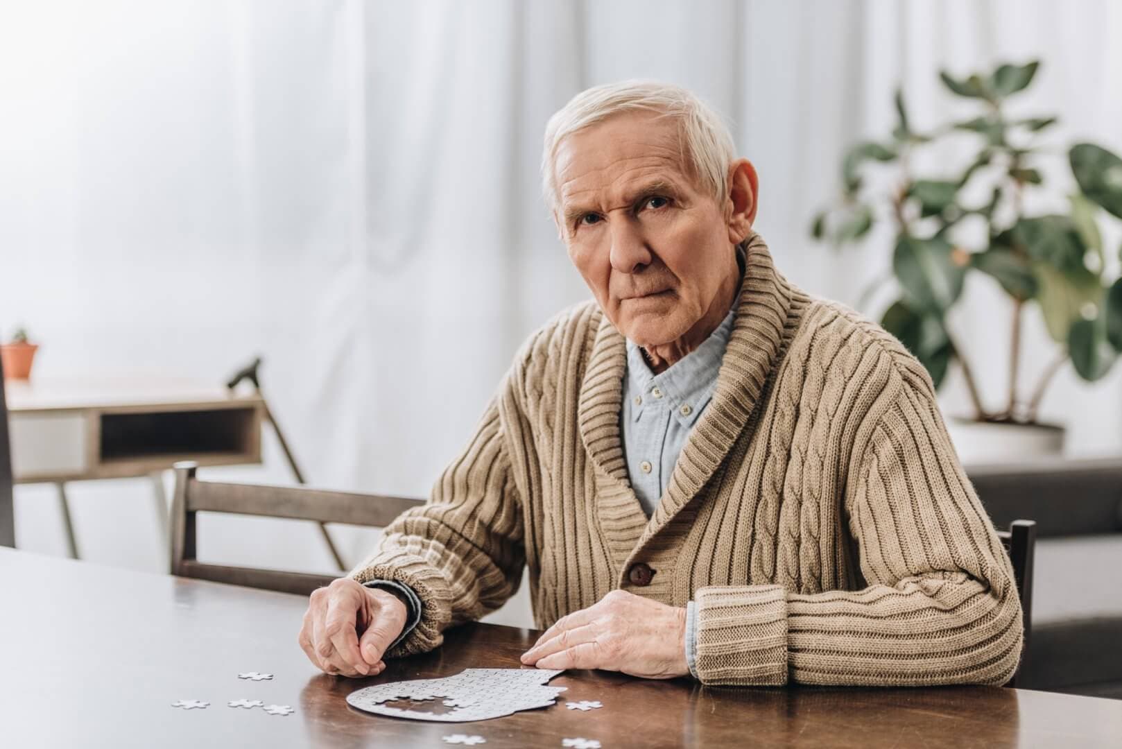 Elderly man in a beige sweater sitting at a table, working on a puzzle, with a pensive expression. - Home Instead