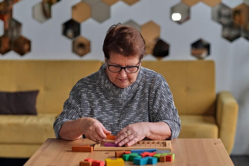 An older woman with glasses focuses on arranging a wooden puzzle at a table in a cozy living room. - Home Instead