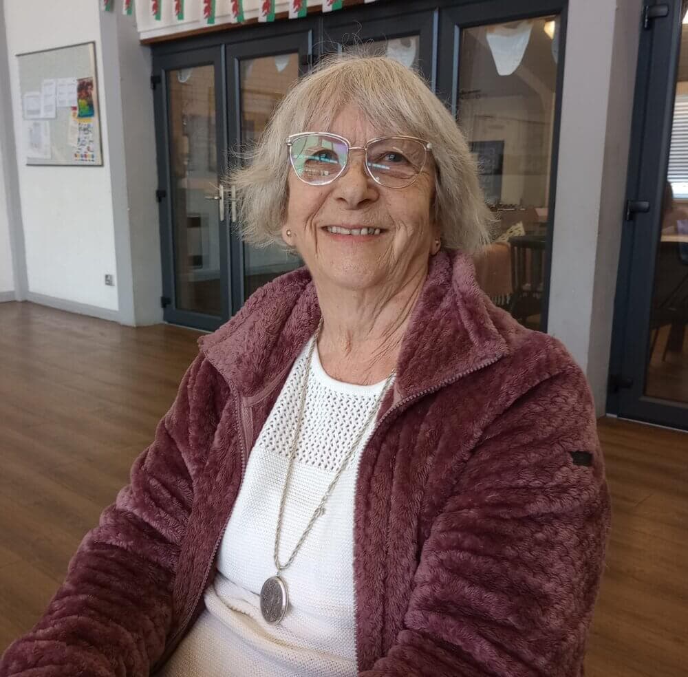 An elderly woman with glasses and a mauve jacket smiles while sitting indoors. Background includes glass doors. - Home Instead