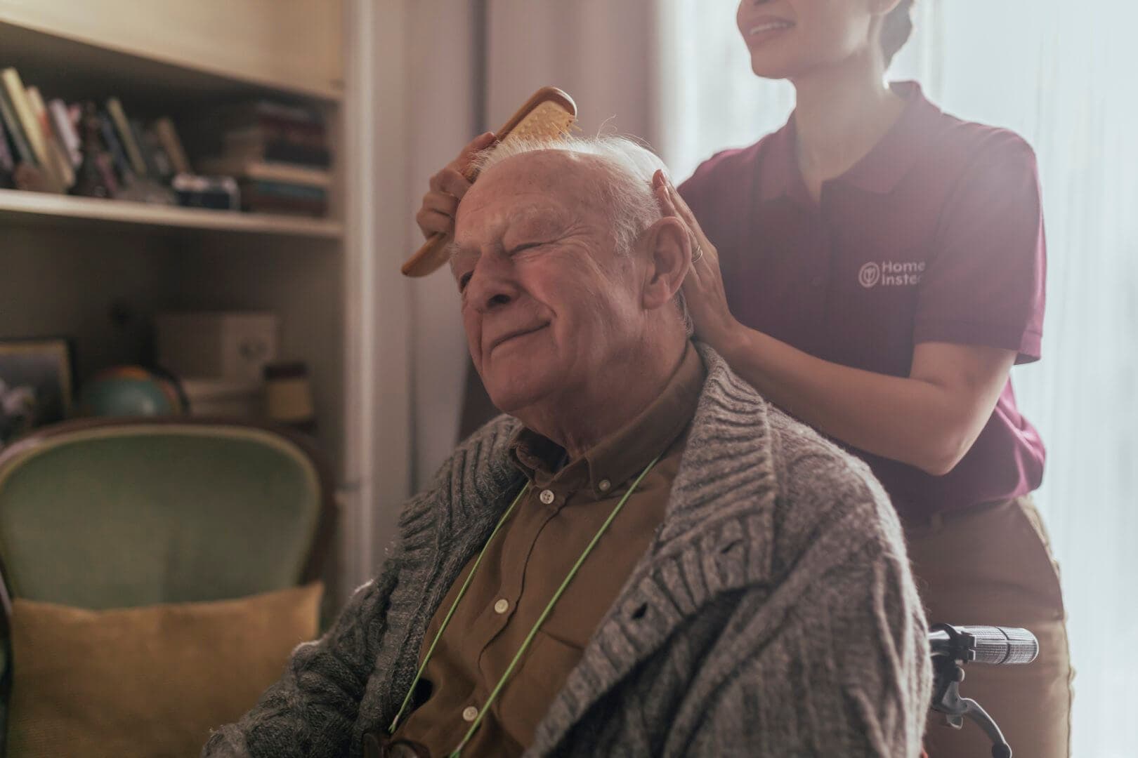 Elderly man in a wheelchair smiling as a caregiver combs his hair in a cozy room. - Home Instead