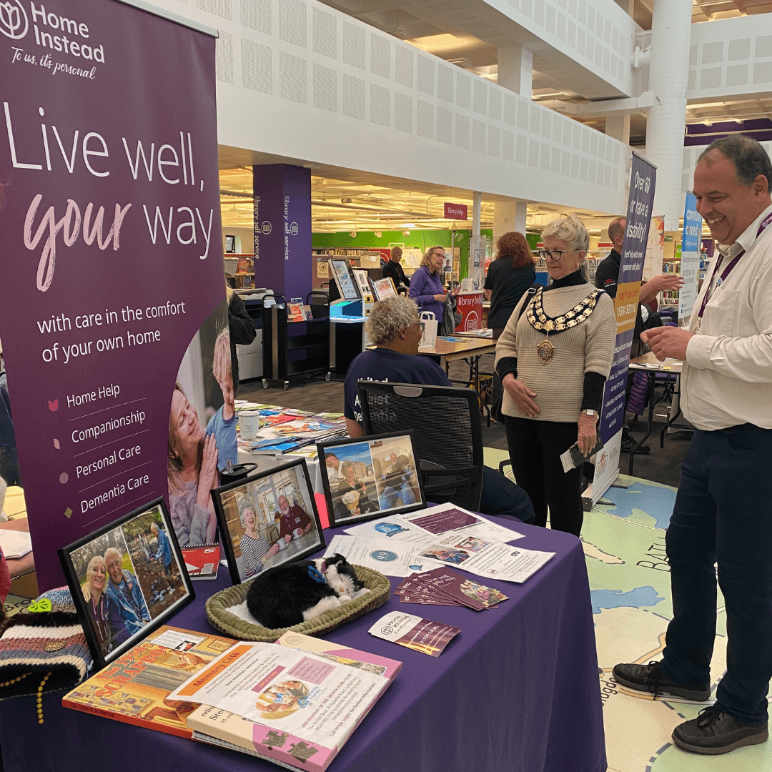 A booth with a "Home Instead" banner, informational materials, and people conversing at a community event. - Home Instead