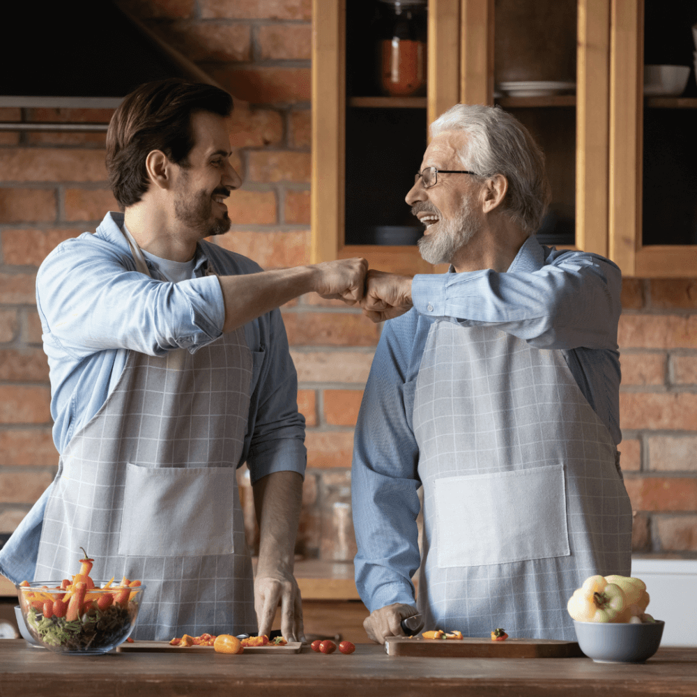 Two men wearing aprons sharing a fist bump while prepping vegetables in a modern kitchen. - Home Instead