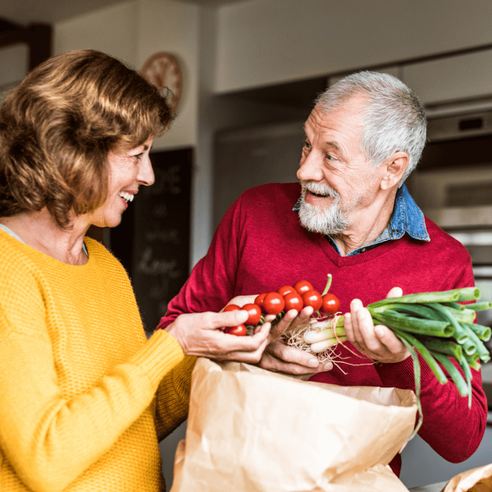 A smiling older couple unpacks groceries in the kitchen, holding tomatoes and green onions from a paper bag. - Home Instead