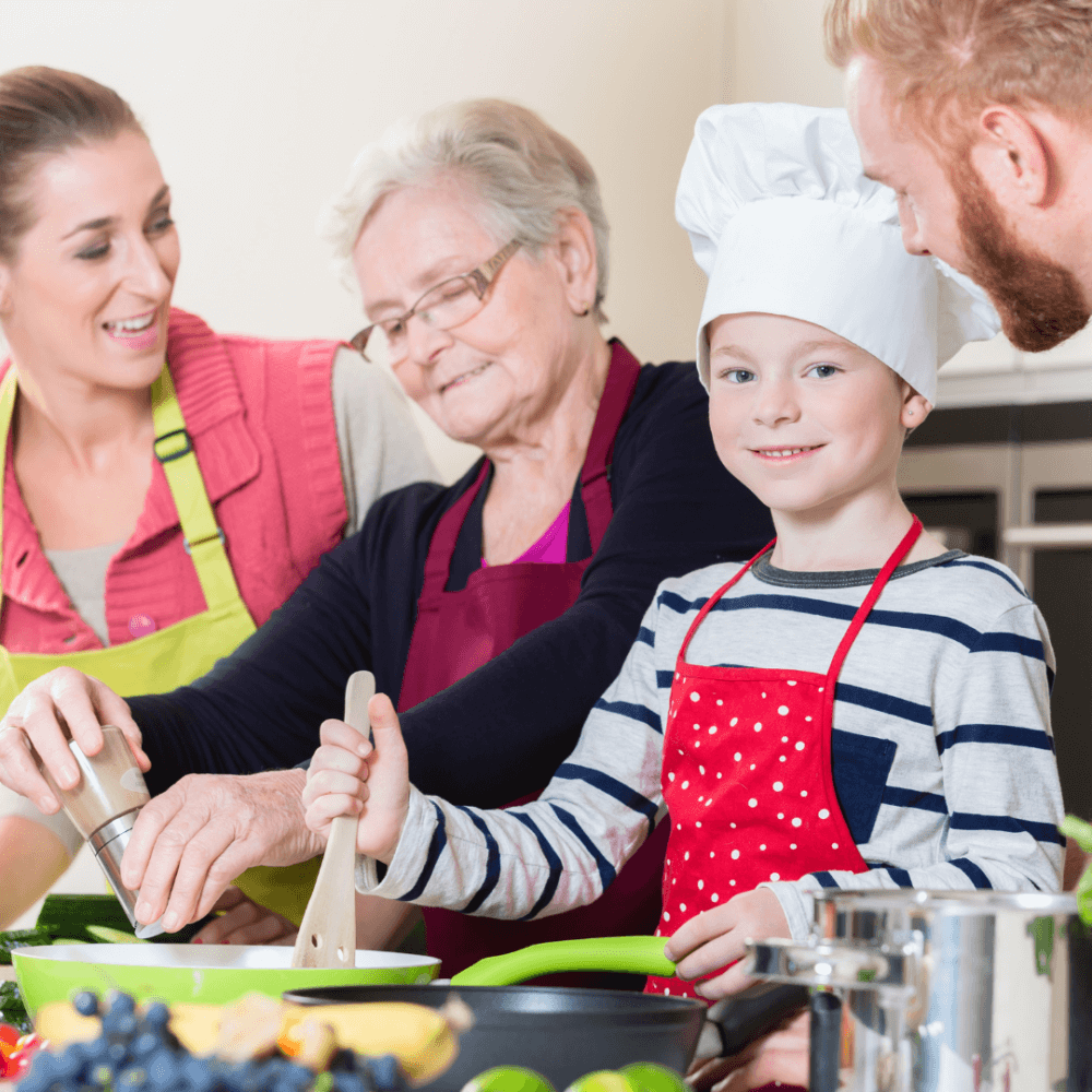 A family in a kitchen, child wearing a chef's hat, adults smiling, and elderly woman seasoning food. - Home Instead