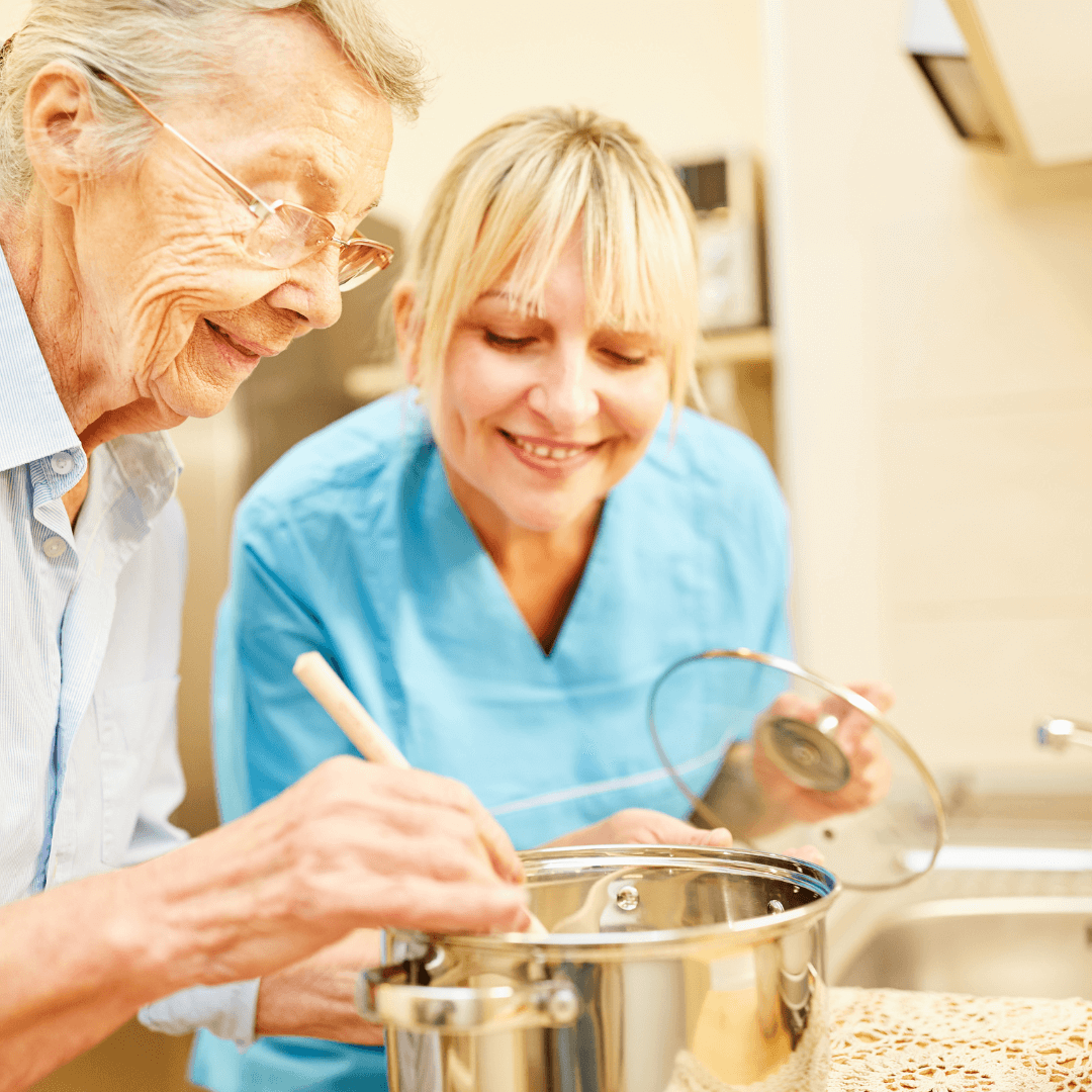 An elderly woman cooking with a wooden spoon in a pot while a nurse or caregiver smiles beside her in a kitchen. - Home Instead