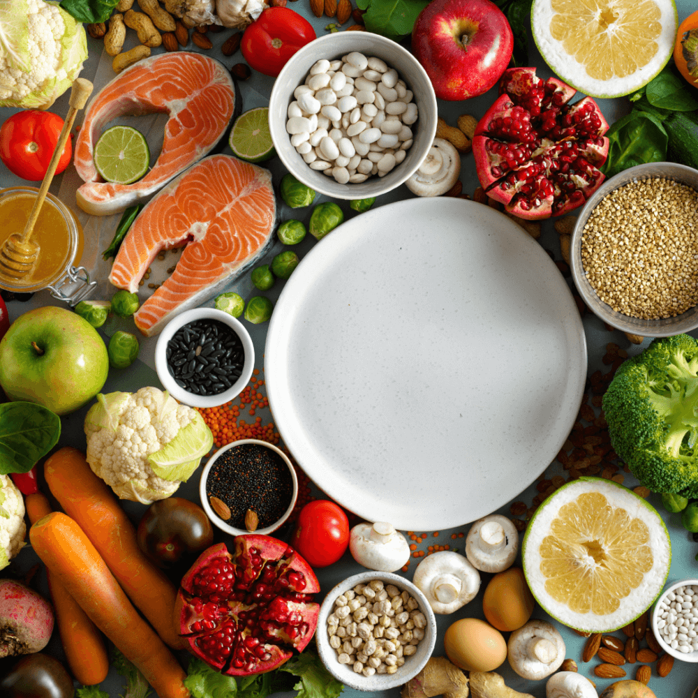 Various fresh foods, including fruits, vegetables, fish, and grains, surrounding an empty white plate on a tabletop. - Home Instead