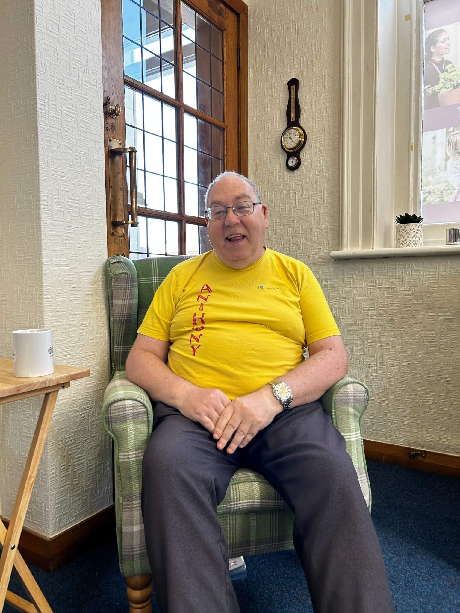 A smiling man in a yellow shirt sits in a green plaid chair next to a small table with a mug on it. - Home Instead