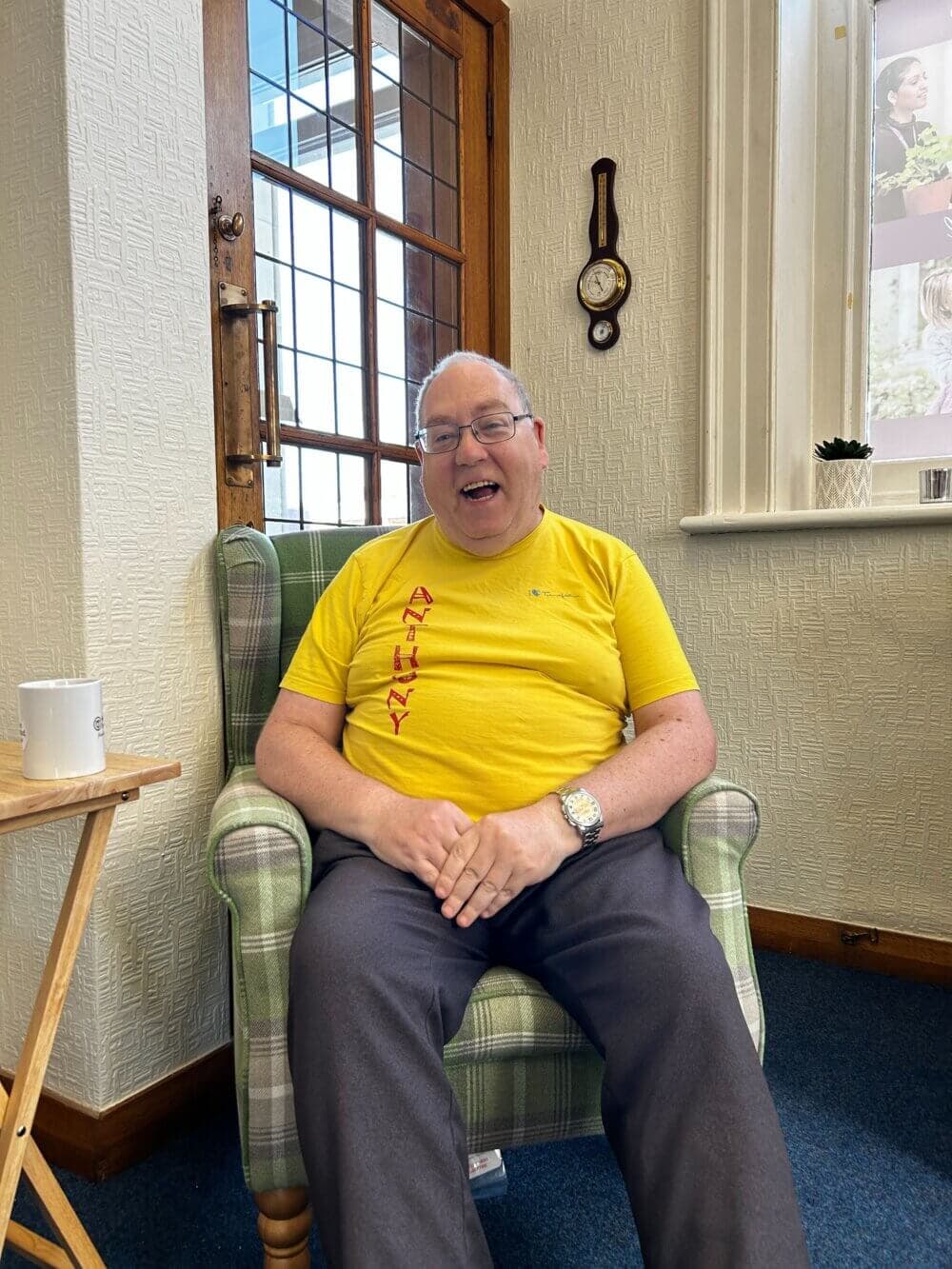 A smiling man in a yellow shirt sits in a plaid armchair in a cozy room. - Home Instead