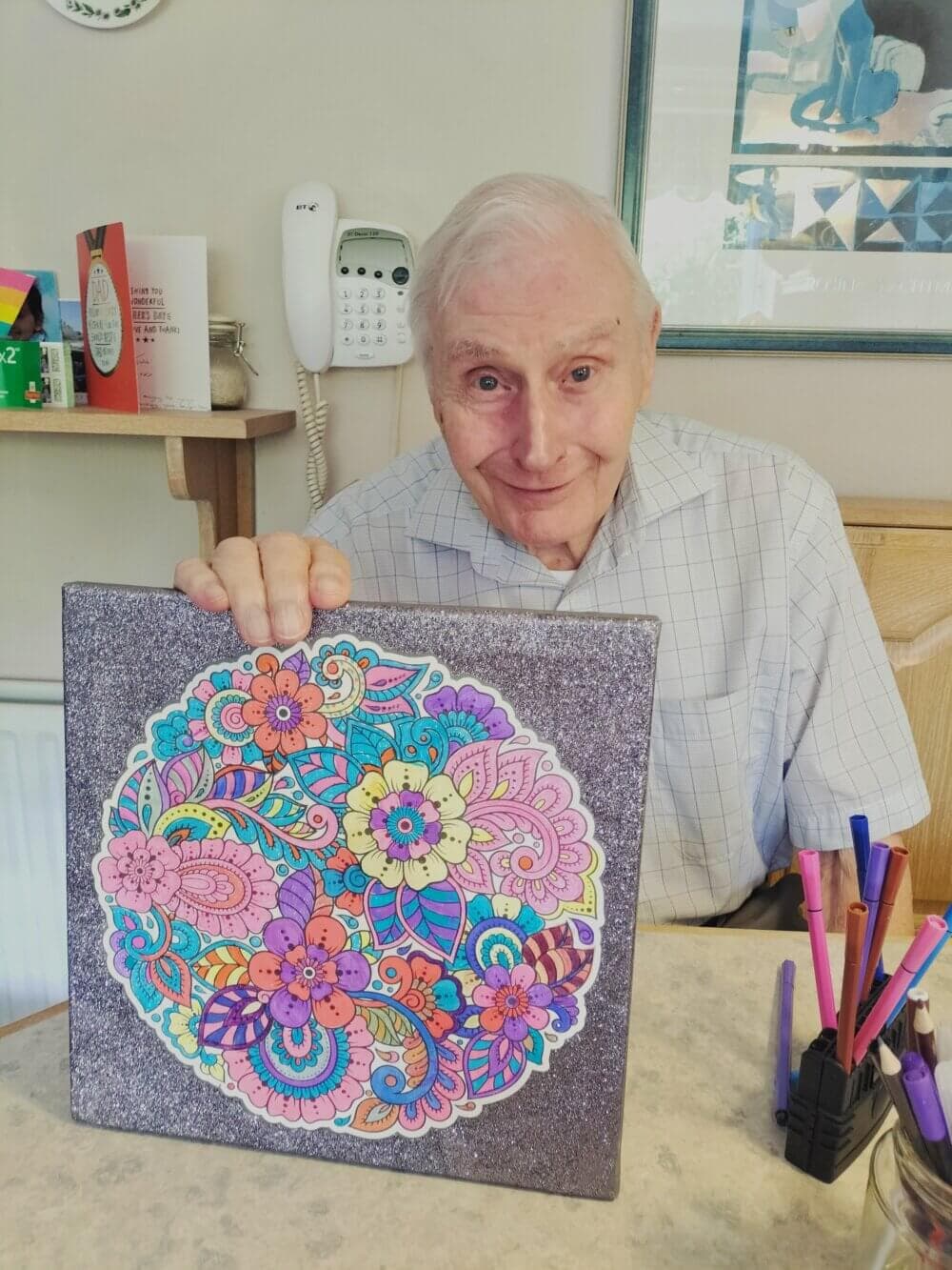 Elderly man proudly holding up a colorful mandala artwork he created, with pens and markers on the table. - Home Instead
