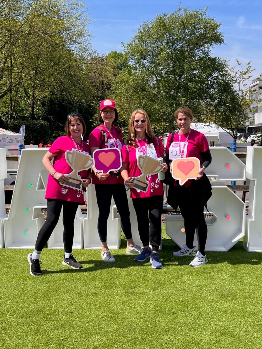 Four women in matching pink shirts holding heart and trophy signs stand in front of a large "RACE" sign outside. - Home Instead