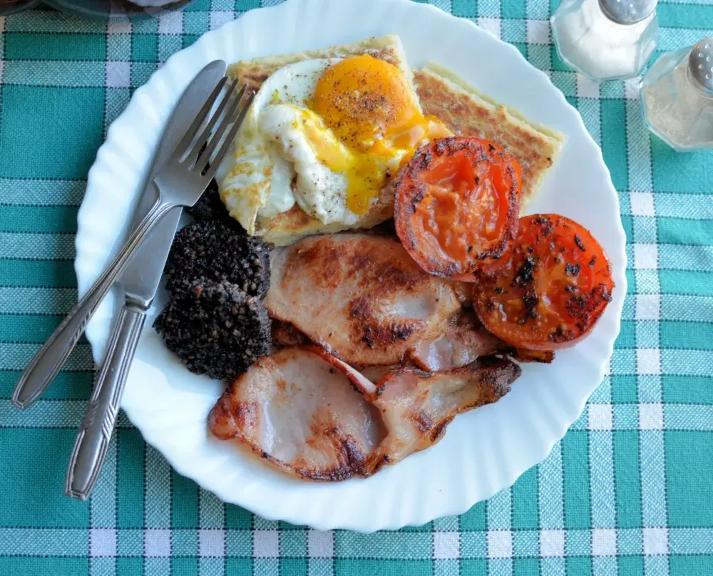 A plate of sunny-side-up eggs, bacon, black pudding, grilled tomatoes, and toast. Silverware is placed on the plate's edge. - Home Instead