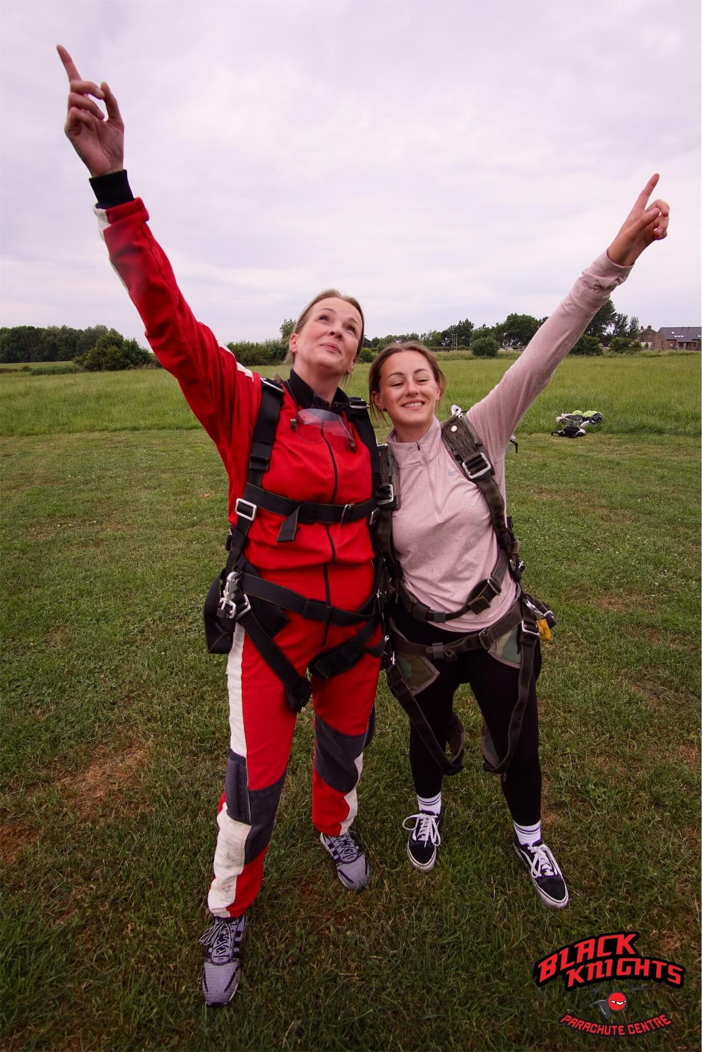 Two people in skydiving gear stand on grass, smiling and pointing upwards. "Black Knights Parachute Centre" logo is visible. - Home Instead