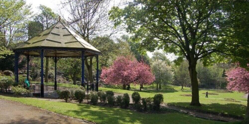 Gazebo in a lush park with pink cherry blossoms and green trees on a sunny day. - Home Instead