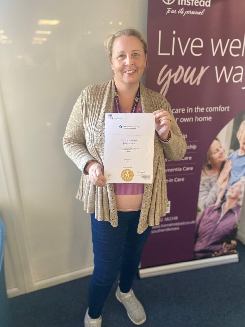 A woman smiles while holding a certificate, standing in front of a banner that says "Live well your way. - Home Instead