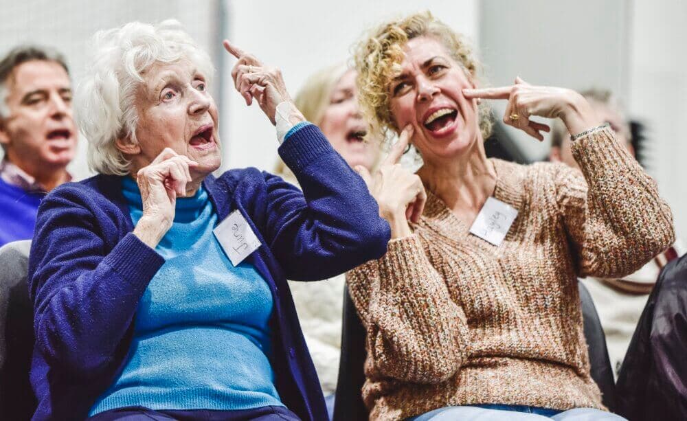 Two women, wearing name tags, animatedly gesturing while sitting in a group with others in the background. - Home Instead