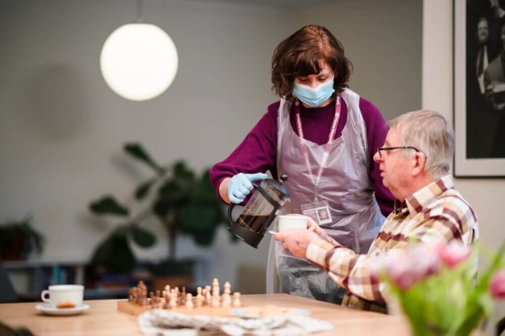 Caregiver wearing mask and gloves pours coffee for an elderly man sitting at a table with a chessboard. - Home Instead