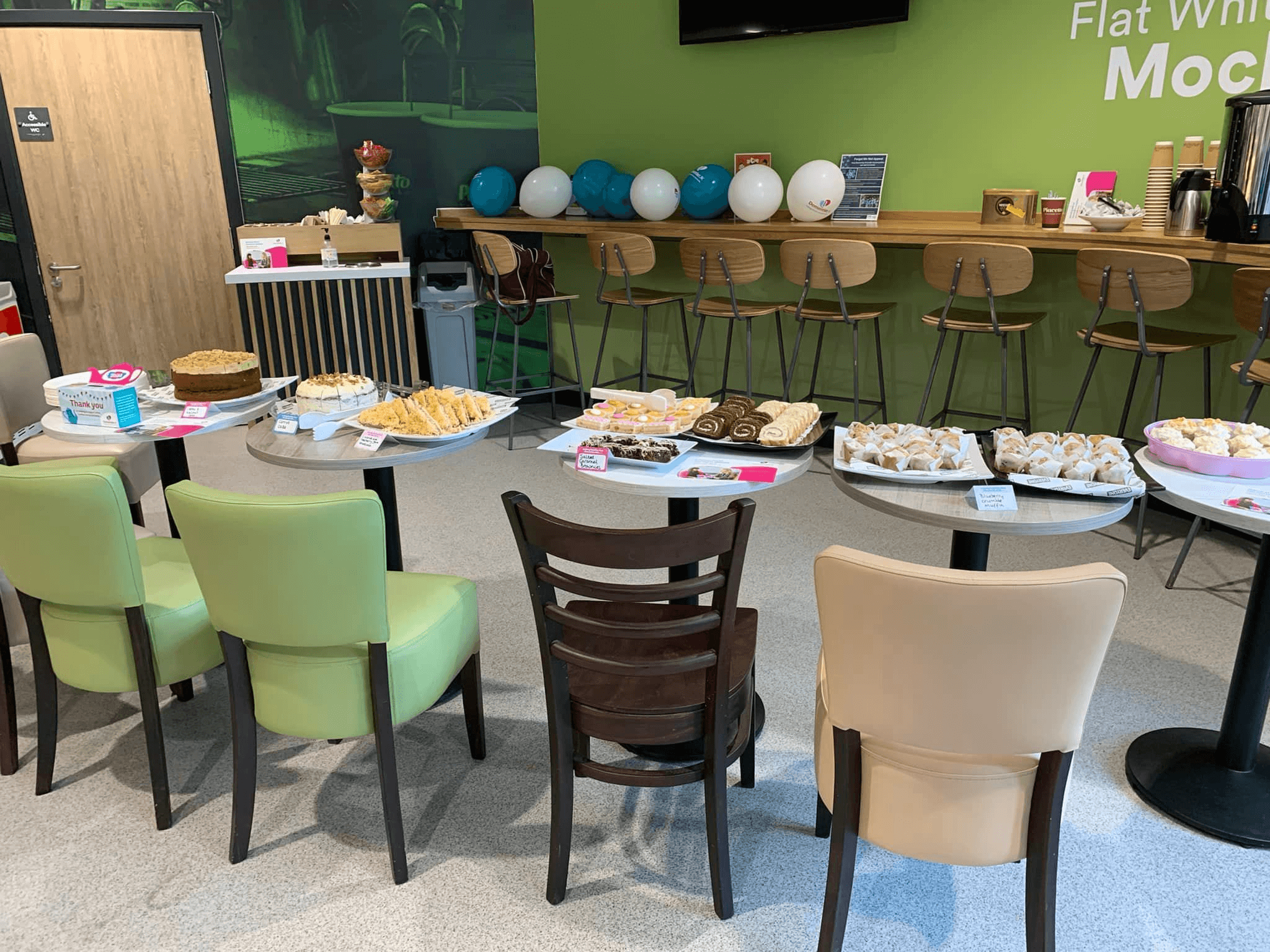 A table with various snacks and chairs arranged in a room, with barstools and a coffee station in the background. - Home Instead