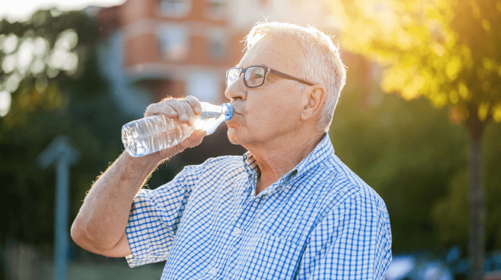 Elderly man with glasses drinking water from a plastic bottle outdoors on a sunny day, wearing a checkered shirt. - Home Instead