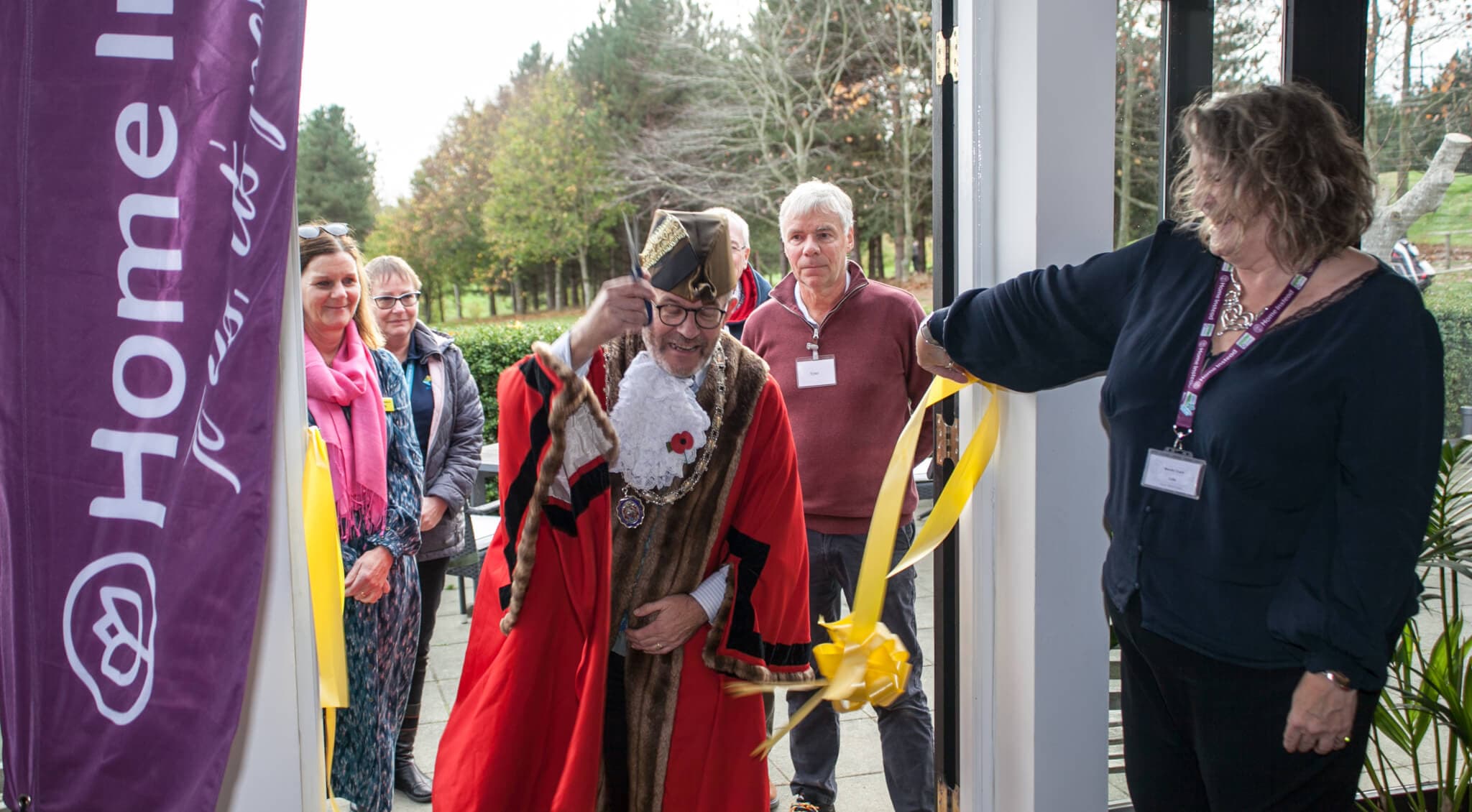 A man in ceremonial attire cuts a yellow ribbon at an opening event, with several people standing nearby. - Home Instead