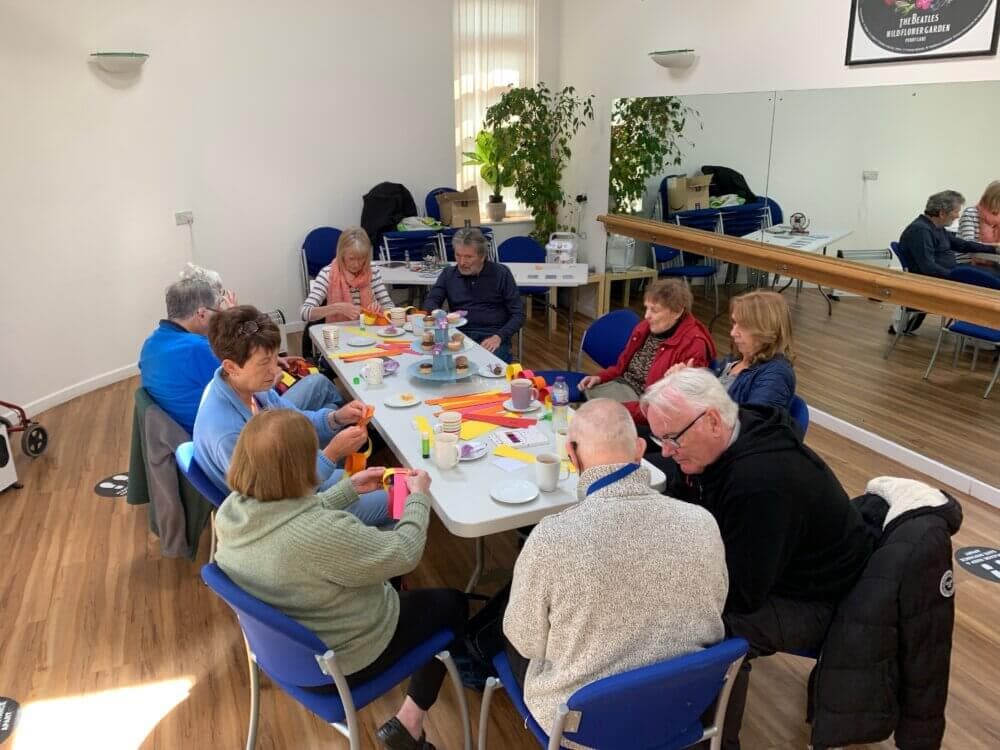 A group of nine senior individuals are sitting around a table, engaged in a crafts activity in a bright room. - Home Instead