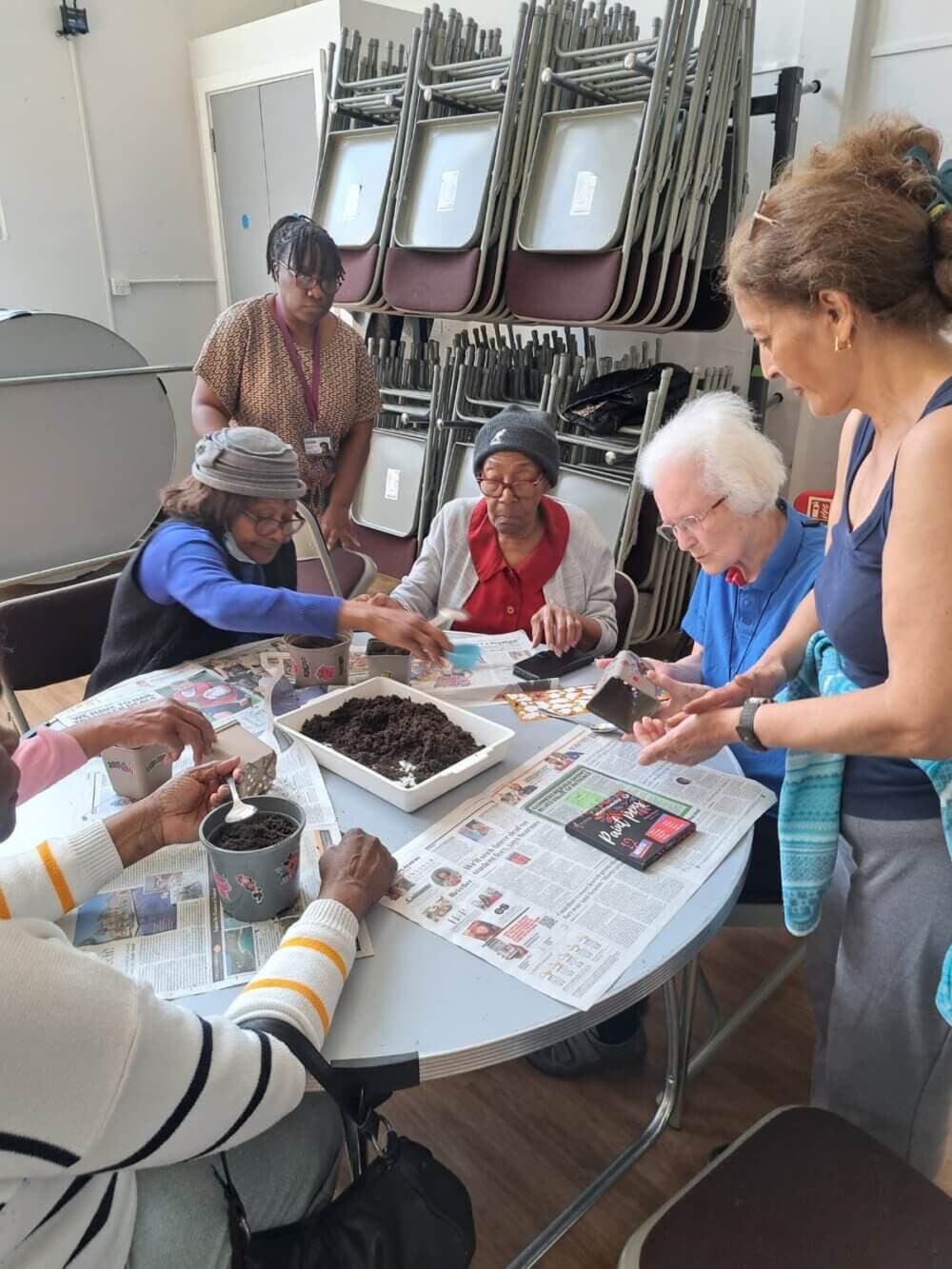 Seniors and a caregiver engage in a group gardening activity, planting seeds at a table covered in newspapers. - Home Instead