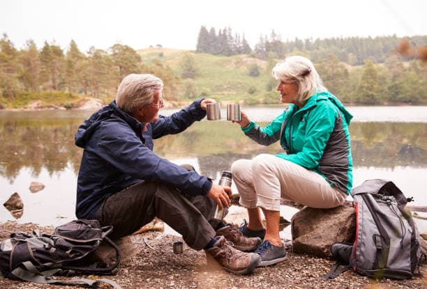 Older couple sitting by a lake, toasting with mugs, and enjoying the outdoors with backpacks nearby. - Home Instead