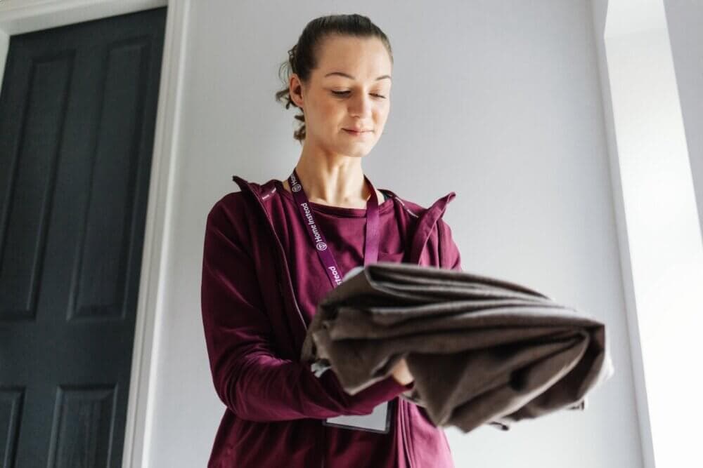 A woman in a maroon outfit folds a cloth while standing near a door in a brightly lit room. - Home Instead
