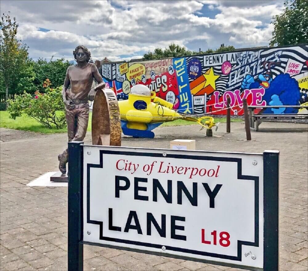 Penny Lane sign with John Lennon statue and Beatles-themed mural in Liverpool, featuring vibrant colors and iconic imagery. - Home Instead