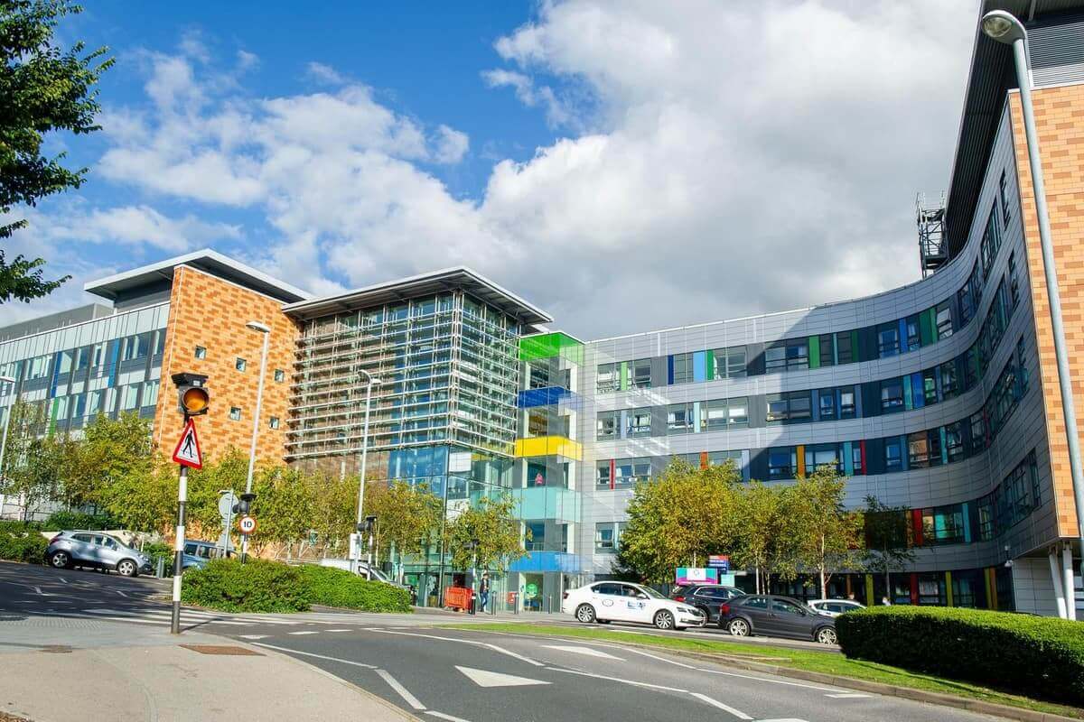 Modern multi-story building with colorful panels, surrounded by trees and parked cars, under a partly cloudy sky. - Home Instead